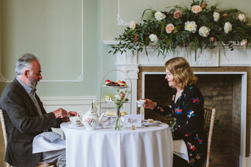 A couple enjoying afternoon tea in the Terrace Room. The tables and mantlepiece are decorated with light pink and white floral arrangements.