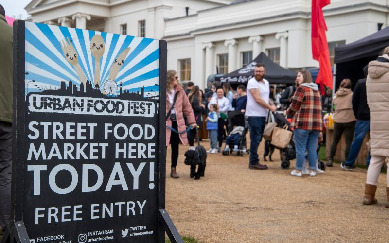 A sign reading 'Street Food Market Here Today' with stalls and a crowd of people in the background.