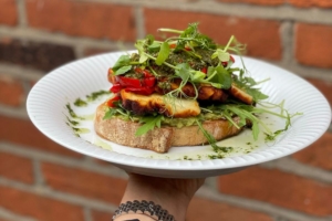 An open-faced sandwich topped with avocado, grilled halloumi, mixed greens and pesto.