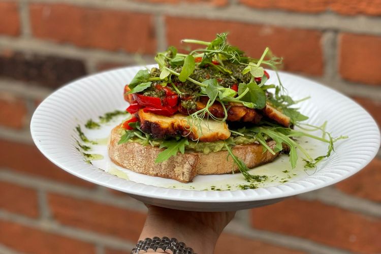 A piece of sourdough toast on a white plate. It is piled high with haloumi, red bell pepper, avocado and greens.