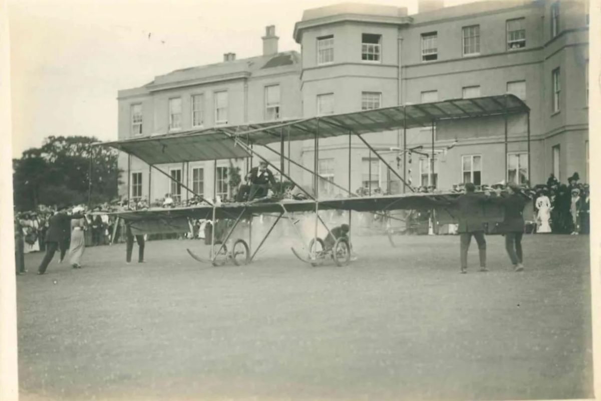 A historical photo of a biplane landing on the back lawn of Hylands House. A large group of spectators are dressed in formal attire.