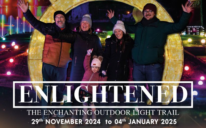 Enlightened 2024. A family wearing warm hats and jackets, smile while standing in front of lit up Christmas decorations.