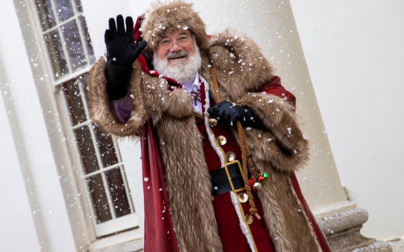 Santa, dressed in a fur-lined red robe, waves to the camera while snow falls around him.