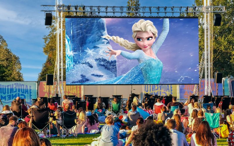 Frozen plays on a large, outdoor cinema screen. The audience is sat on a variety of lawn chairs and blankets.