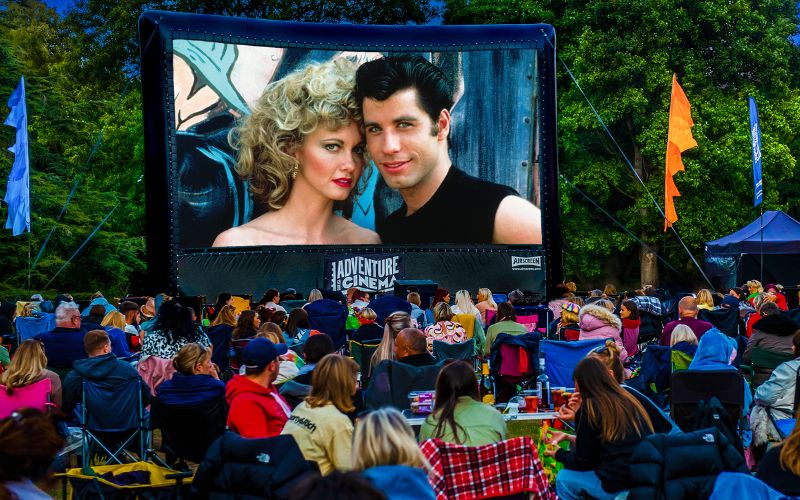 Grease plays on a large, outdoor cinema screen. The audience is sat on a variety of lawn chairs and blankets.