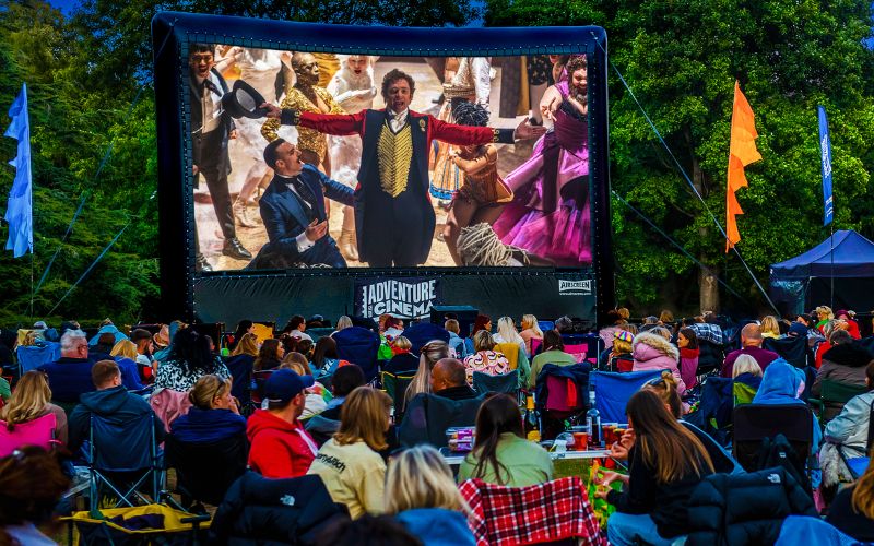 The Greatest Showman plays on a large, outdoor cinema screen. The audience is sat on a variety of lawn chairs and blankets.