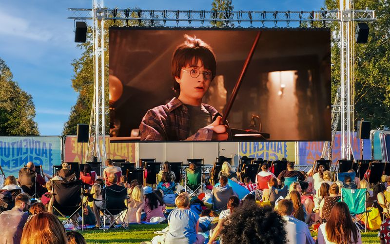 Harry Potter plays on a large, outdoor cinema screen. The audience is sat on a variety of lawn chairs and blankets.