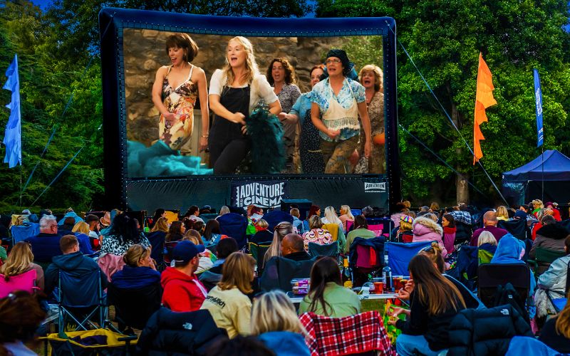 Mamma Mia plays on a large, outdoor cinema screen. The audience is sat on a variety of lawn chairs and blankets.