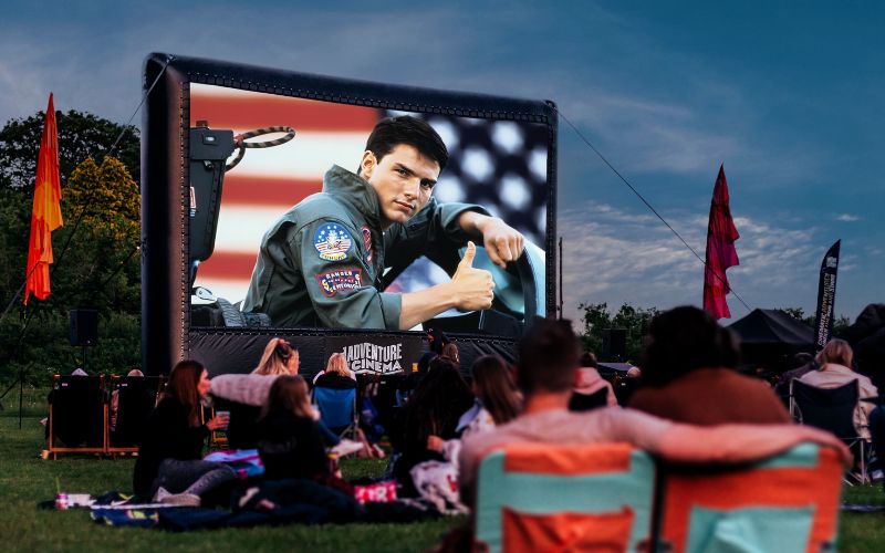 Top Gun plays on a large, outdoor cinema screen. The audience is sat on a variety of lawn chairs and blankets.