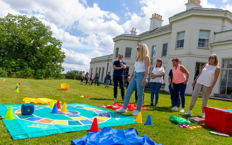 A blonde woman stands in front of a team of people on the back lawn with Hylands House in the background. She stands in front of a ground based board game.