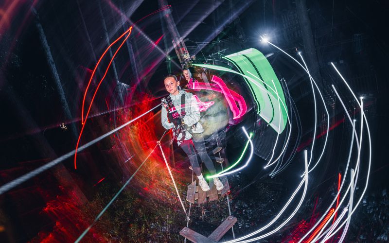 Children walking across a high ropes course at night with fun lighting.
